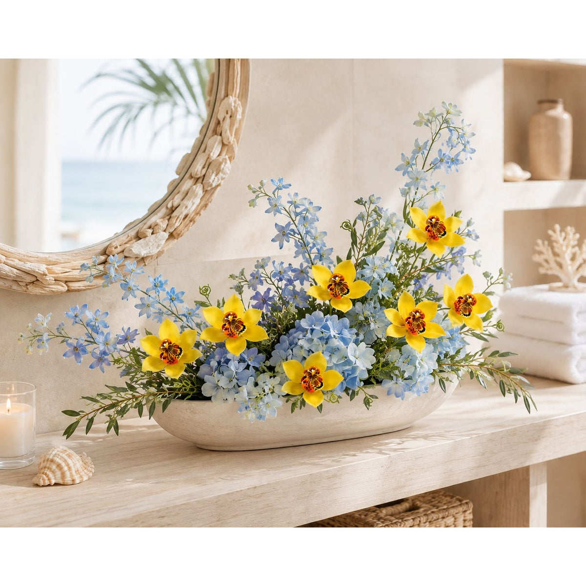 A decorative floral arrangement in a white oval bowl on a light wood console table, featuring bright yellow flowers, light blue flowers, and green foliage, styled in a bright beach-inspired room with a mirror, candle, and folded towels.