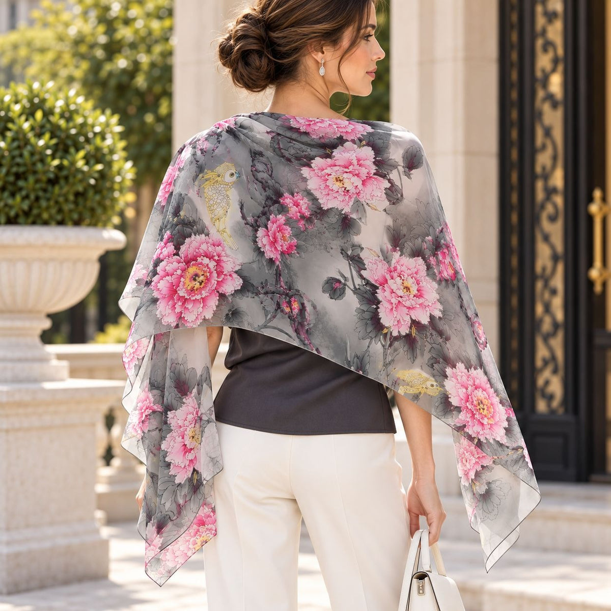 A woman shown from the back wearing a sheer gray shawl with a large pink floral print, styled over a black top and cream trousers in an outdoor architectural setting.