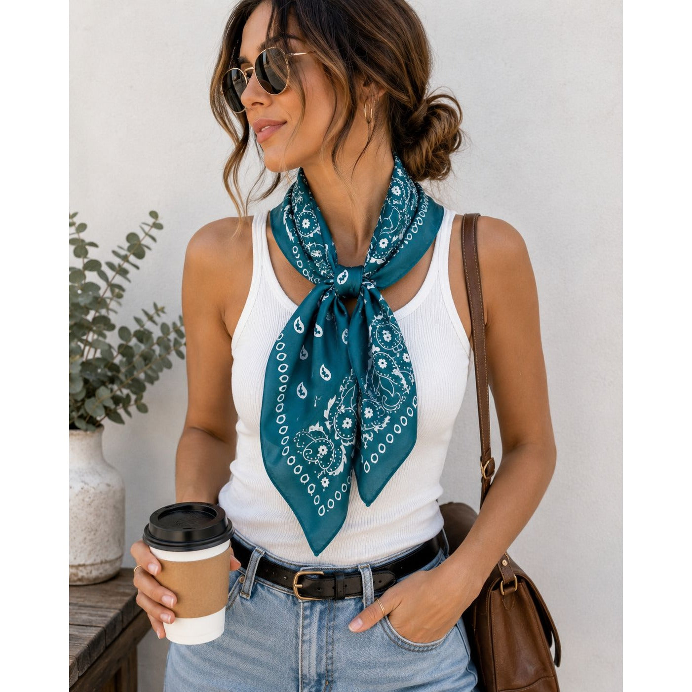 A woman wearing a teal paisley bandana scarf tied around her neck, styled with a white ribbed tank top, blue jeans, sunglasses, and a brown shoulder bag, holding a coffee cup.