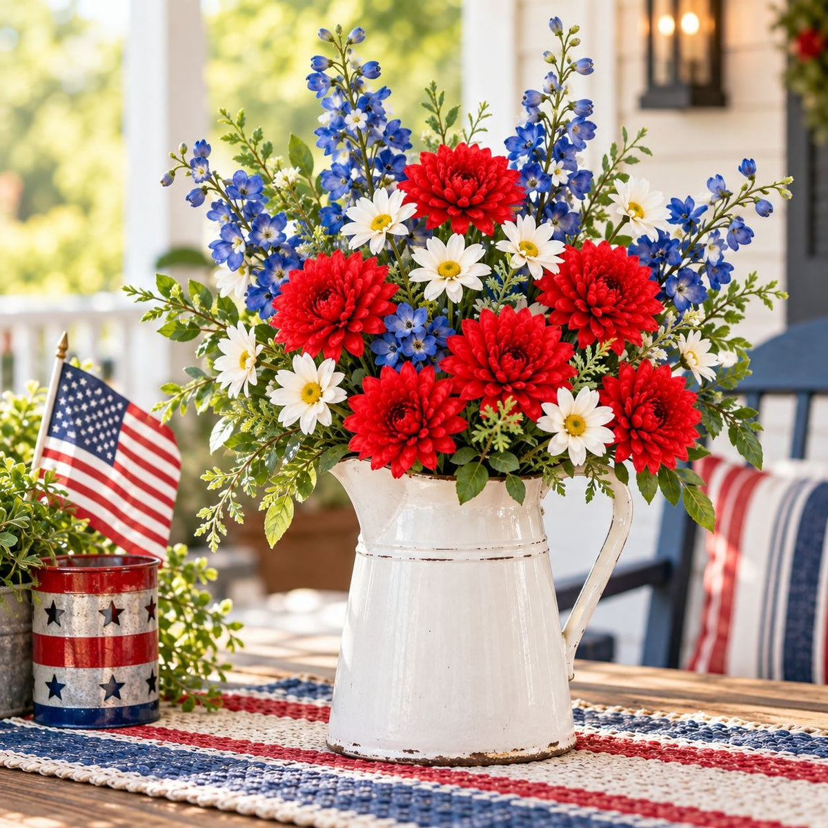 A patriotic floral arrangement in a white metal pitcher, featuring red, white, and blue flowers with green foliage, displayed on a porch table next to a small American flag can.
