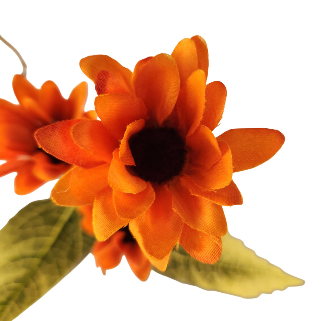 Bright orange fabric sunflower artificial flowers with dark brown centers and green leaves on a white background.