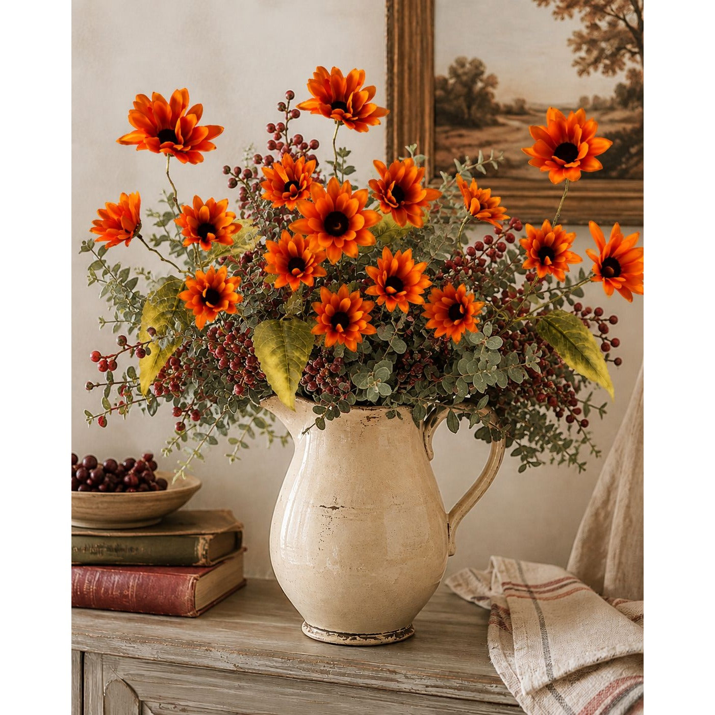 A large cream-colored ceramic pitcher vase filled with a dense arrangement of orange daisy-like flowers, green foliage, and small dark berries, displayed on a rustic wooden table in a styled interior setting.