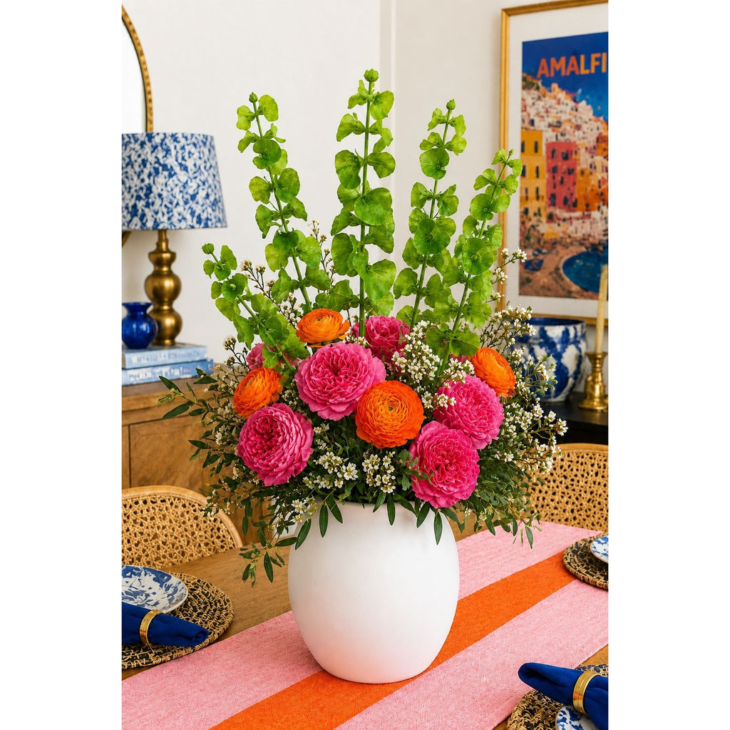 A vibrant artificial flower arrangement in a white ceramic vase, featuring bright pink and orange flowers with green foliage, set on a pink and orange striped tablecloth in a styled dining room.