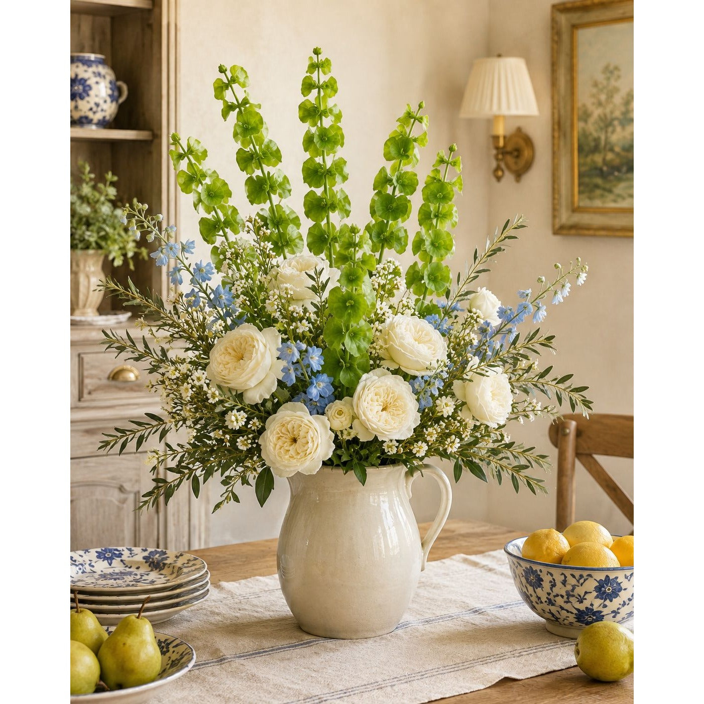 A large floral arrangement in a cream ceramic pitcher vase, featuring white roses, blue flowers, and green foliage, displayed on a dining table in a warm, traditional room.