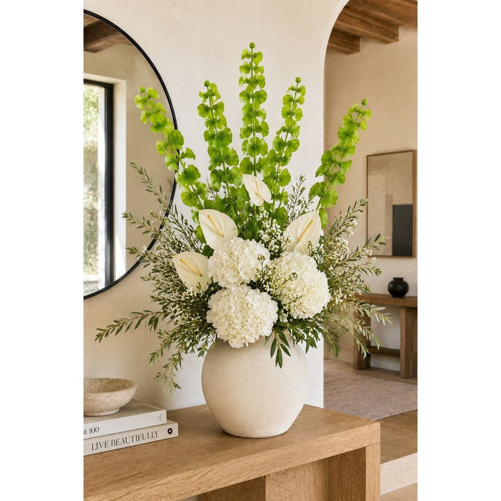A large floral arrangement in a round beige ceramic vase, featuring white lilies, white hydrangeas, and tall green snapdragons, displayed on a wooden console table in a modern, warm-toned interior.