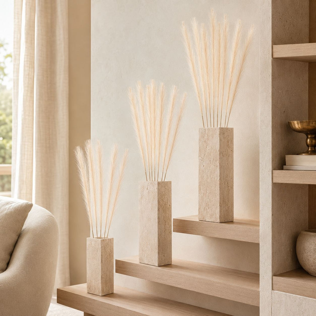 Three rectangular stone vases filled with tall dried pampas grass arranged on light wood steps in a softly lit interior setting.