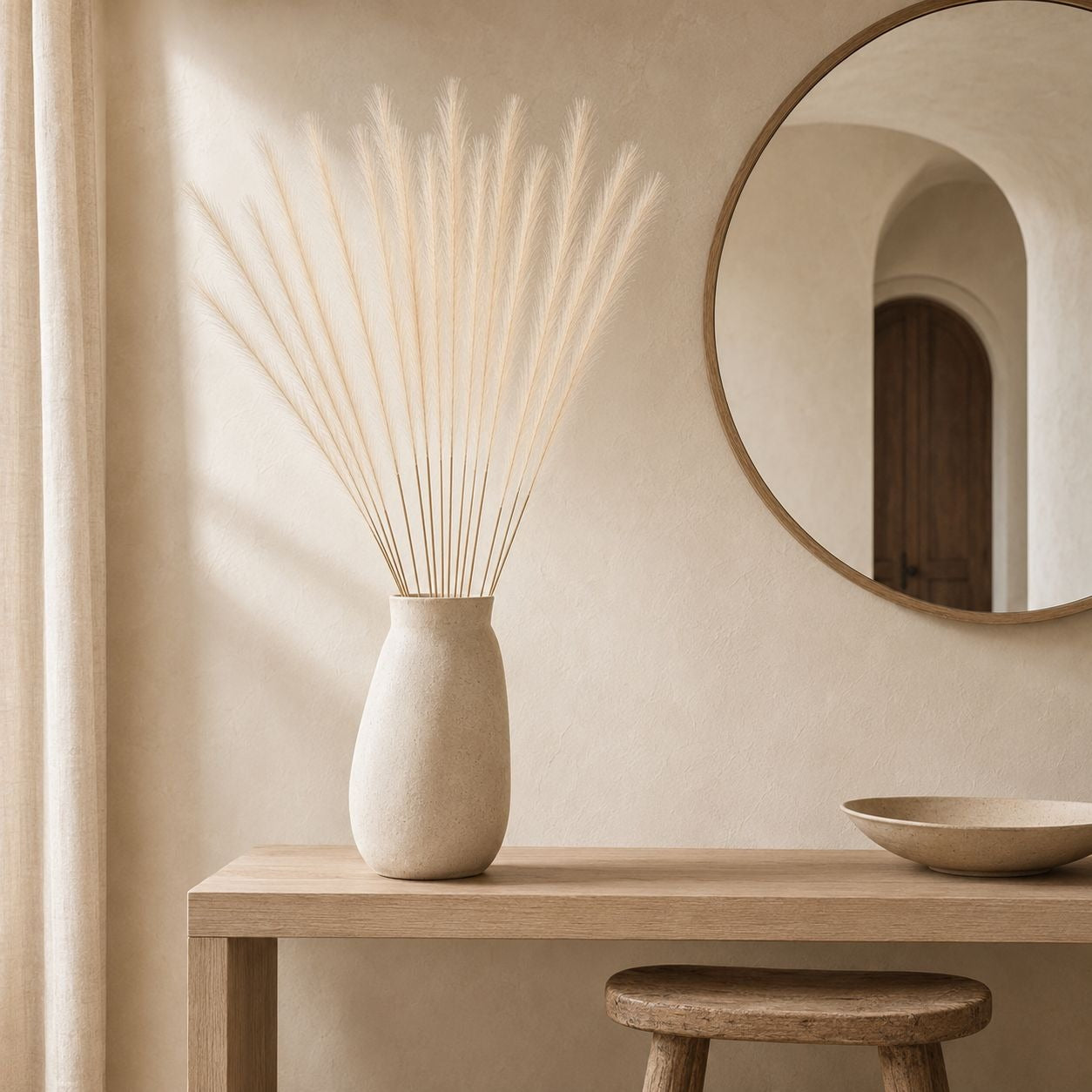 A beige ceramic vase holding dried pampas grass, styled on a light wood console table in a warm neutral interior with a round mirror and a small bowl.