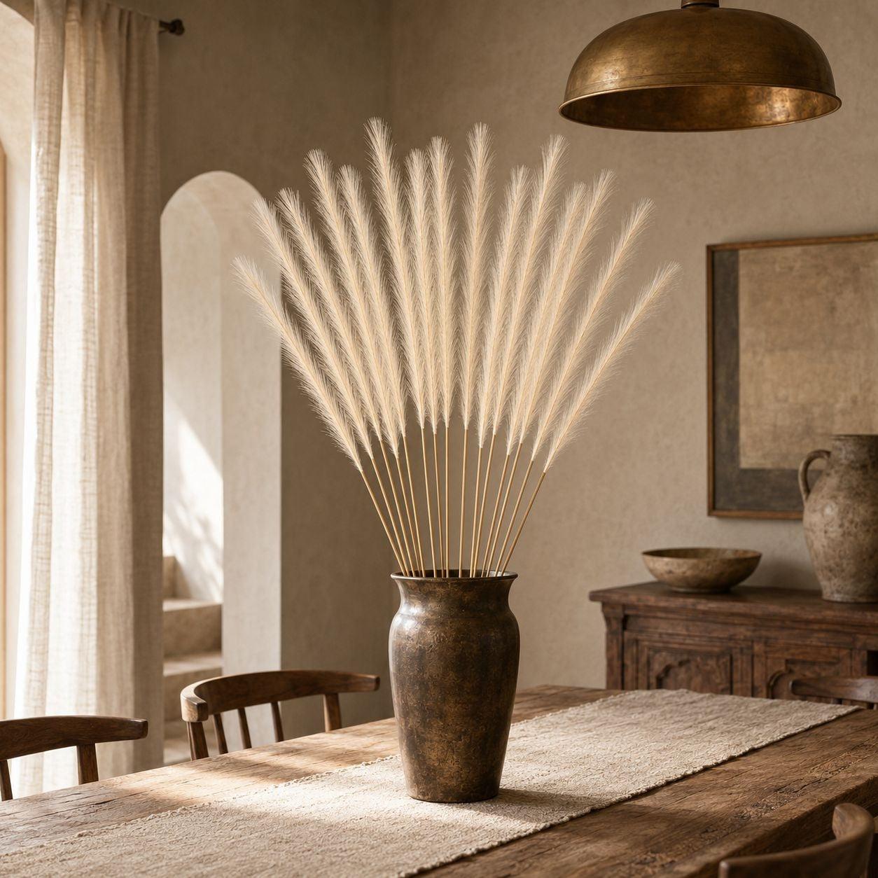 A tall rustic brown ceramic vase filled with a large arrangement of fluffy cream pampas grass, placed on a wooden dining table in a warm, neutral-toned room.