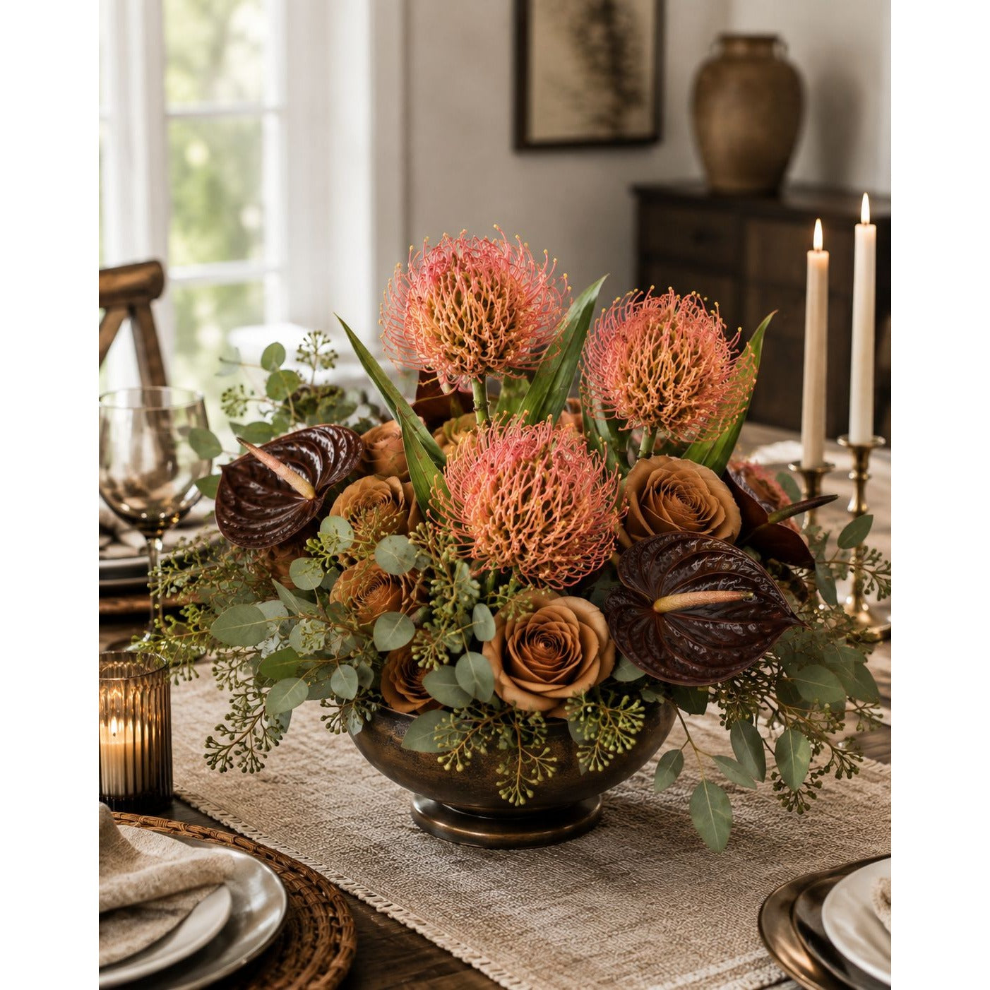 A floral centerpiece arrangement in a dark bronze bowl on a dining table, featuring large coral-pink protea-like blooms, tan and dark brown roses, anthuriums, and green foliage, styled in a warmly lit home dining room.