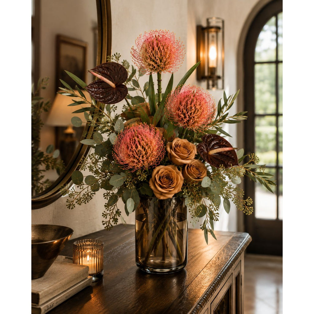 A floral arrangement in a dark glass vase on a wooden console table, featuring coral protea-like blooms, tan roses, green foliage, and dark anthuriums, set in a warmly lit interior with a mirror and sconce.