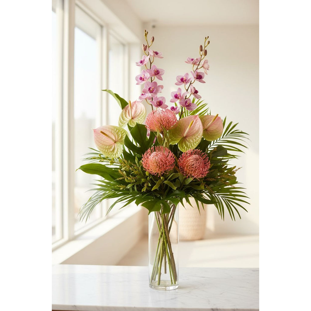 A vibrant floral arrangement featuring pink orchids, pink protea-like flowers, and green anthuriums with palm fronds, presented in a clear glass vase on a light marble surface near a bright window.