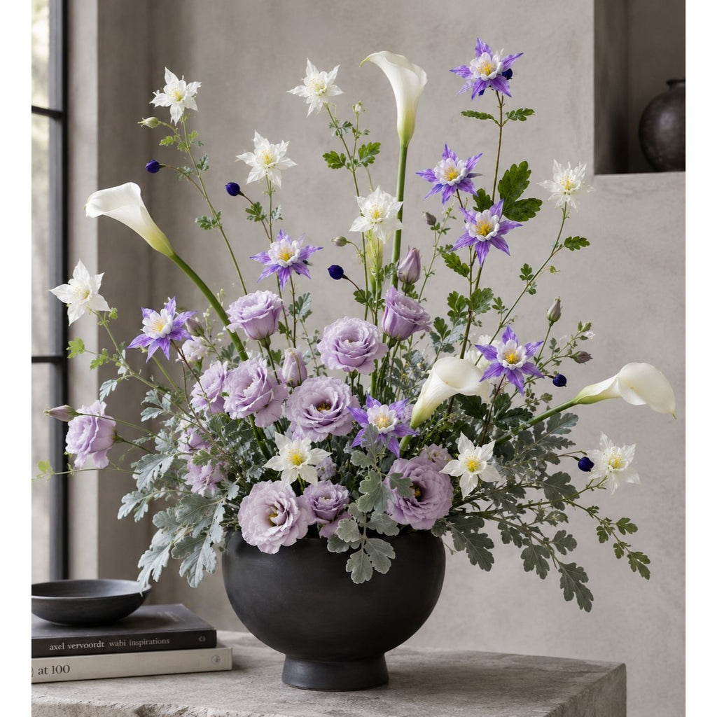 A large floral arrangement in a matte black round vase, featuring lavender and white flowers with green foliage, displayed on a stone table in a modern interior setting.
