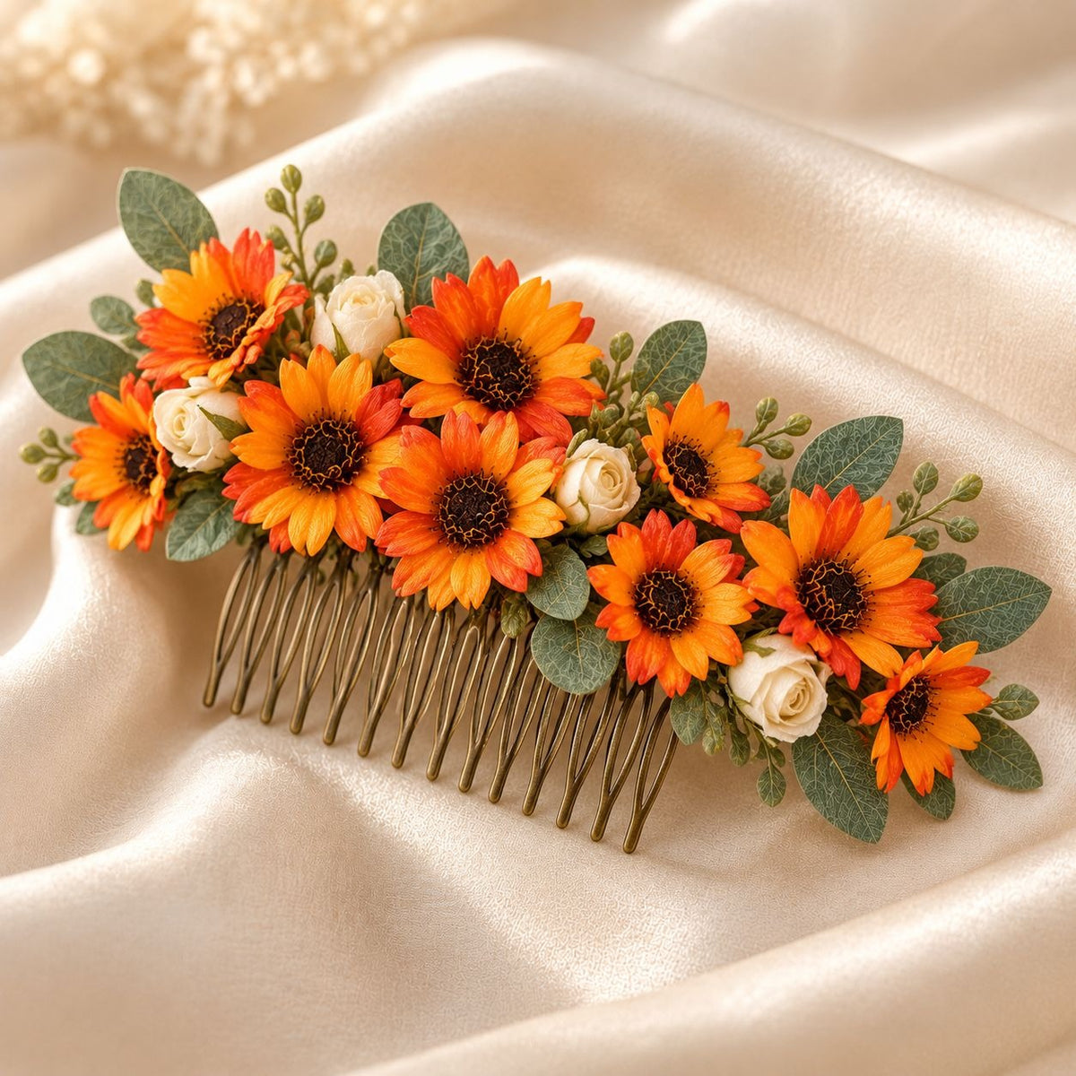 A floral hair comb accessory with orange and yellow sunflowers, small white roses, green leaves, and a gold-toned metal comb base, displayed on draped cream satin fabric.