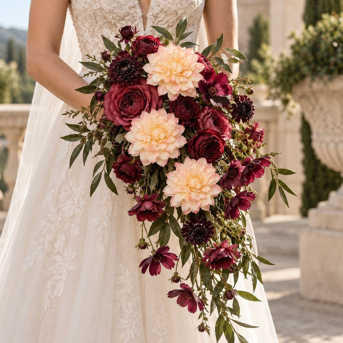 A bridal bouquet with deep burgundy roses, pale blush peonies, and green foliage, held by a bride in a white lace wedding dress in an outdoor garden setting.