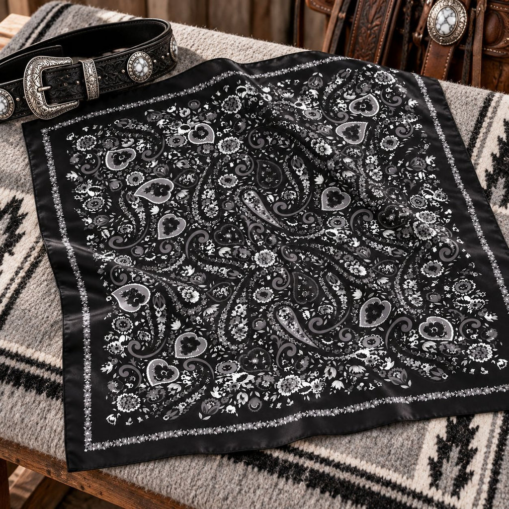 A black bandana with an intricate silver paisley and floral pattern, styled on a gray and black patterned western blanket next to a black leather belt with silver conchos.