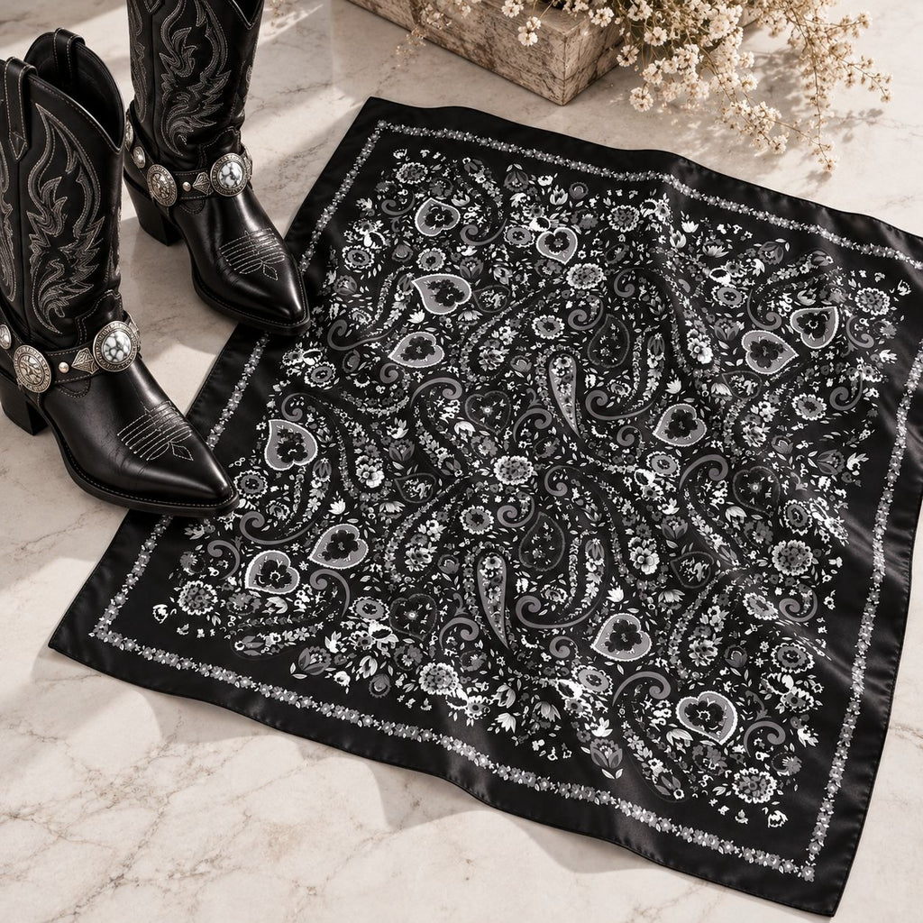 A black bandana with a detailed silver paisley and floral pattern, styled on a light marble surface next to a pair of black western boots with silver conchos.