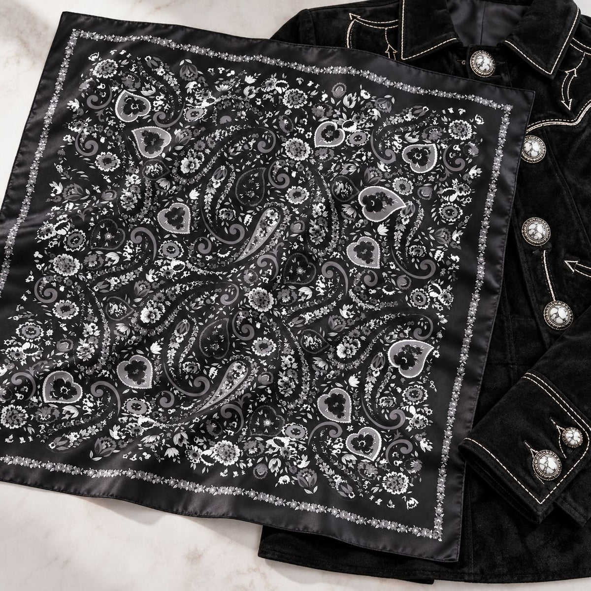 A black satin bandana with an intricate silver paisley and floral pattern, styled flat on a white marble surface next to a black velvet jacket with silver buttons.