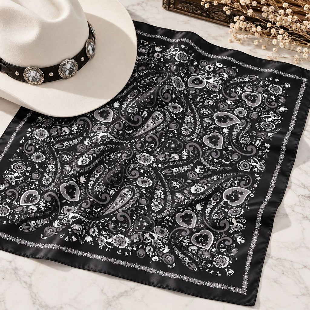 A black satin bandana with a white paisley pattern, styled on a white marble surface next to a white cowboy hat with a black leather and silver concho strap.