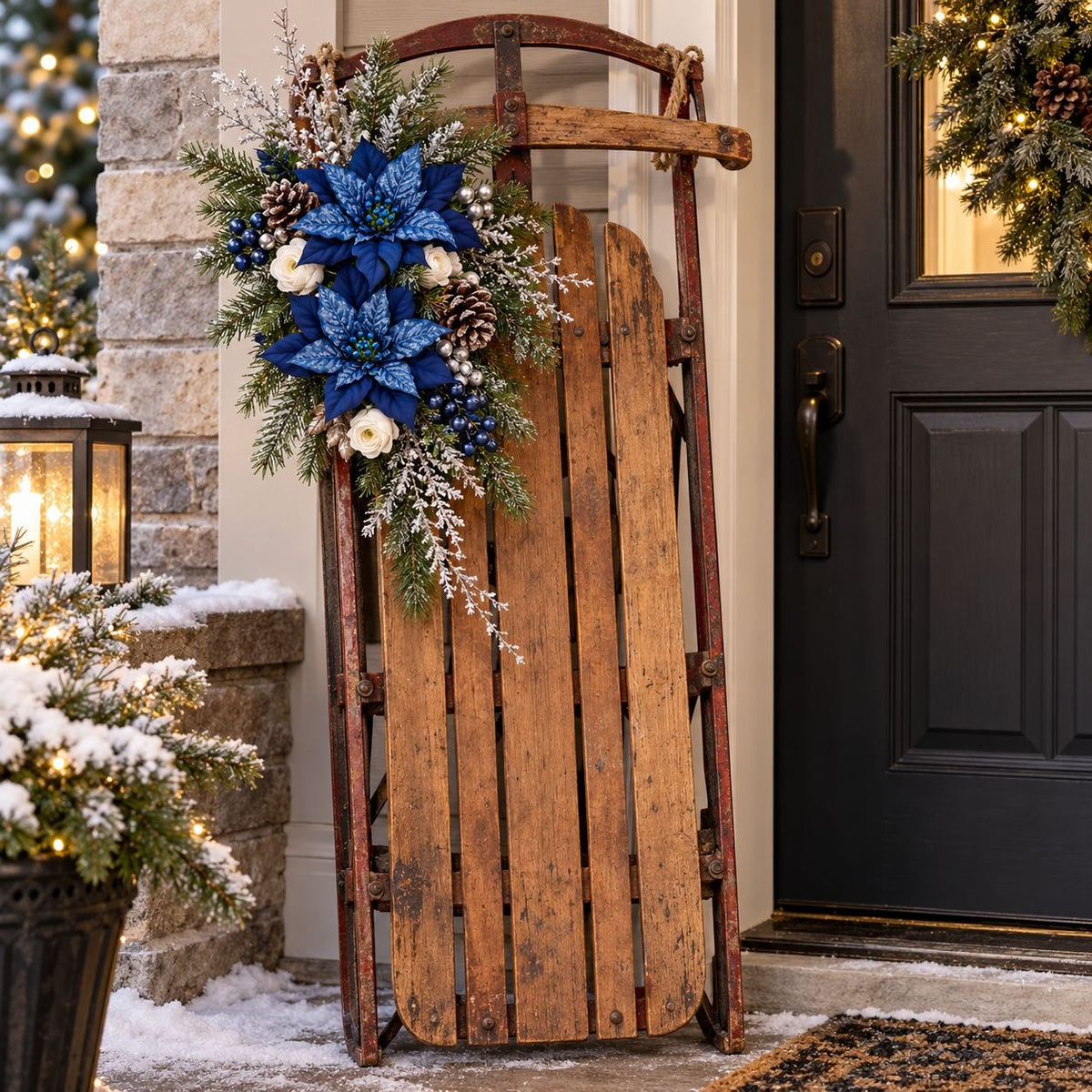 A rustic wooden sled decorated with a large blue and white poinsettia floral arrangement, set on a snowy front porch next to a dark door and stone wall.