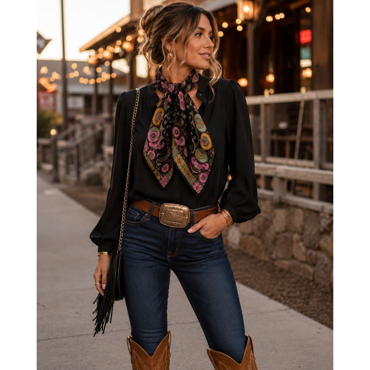A woman wearing a black long-sleeve blouse, blue jeans, and brown cowboy boots, accessorized with a black and multicolor paisley patterned scarf, a black fringe handbag, and a large western-style belt buckle, standing outdoors in front of a rustic building at dusk.