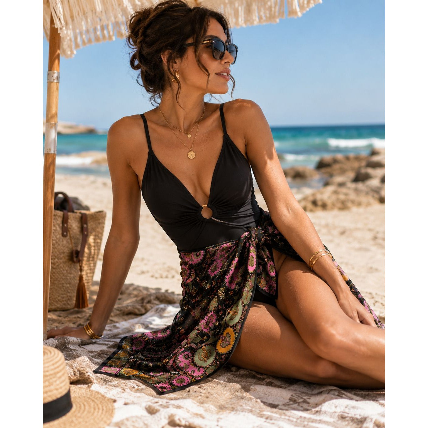 A woman sits on a sandy beach under a woven umbrella, wearing a black halter-style one-piece swimsuit with a deep V-neck and a floral sarong wrap. The background shows the ocean, rocks, and blue sky.