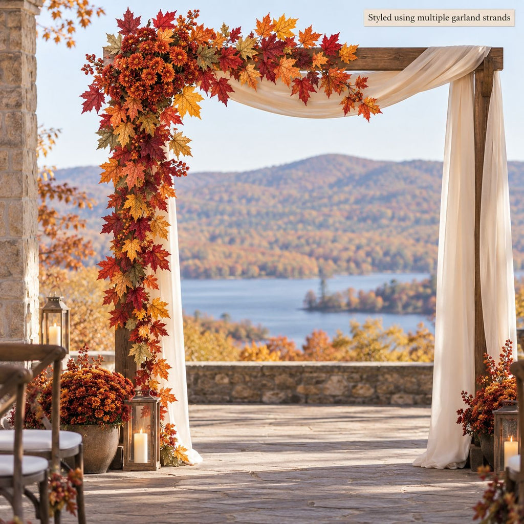An autumn-themed wedding arch decorated with red, orange, and yellow artificial maple leaves and cream draping fabric, set on a stone patio overlooking a scenic lake and forest landscape.