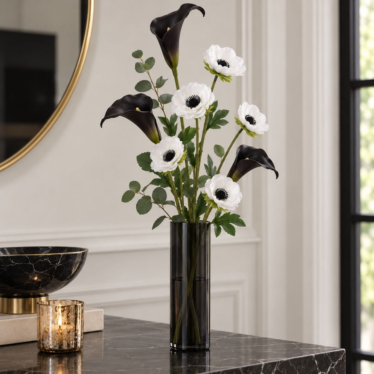 An artificial floral arrangement featuring black calla lilies and white anemone flowers with green leaves, displayed in a tall black glass vase on a dark marble console table.