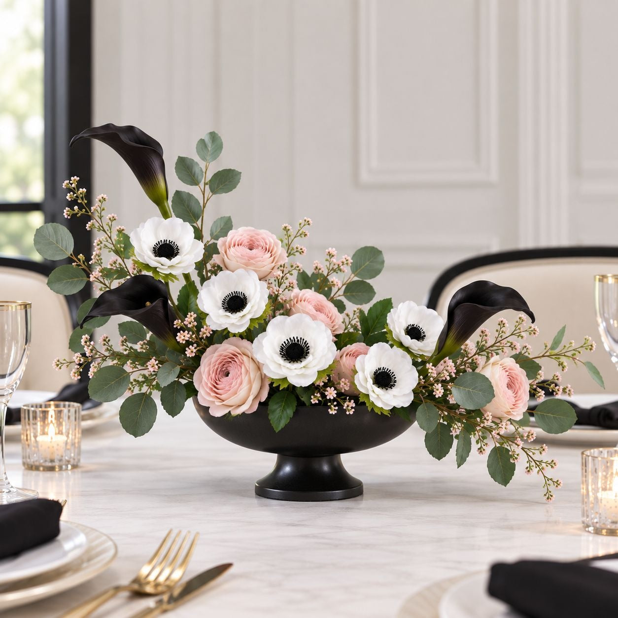 A floral centerpiece arranged in a black pedestal bowl on a dining table, featuring black calla lilies, white anemone flowers with black centers, blush pink roses, green eucalyptus leaves, and small pale pink filler blossoms. The setting includes tableware, candles, chairs, and a softly lit interior background.