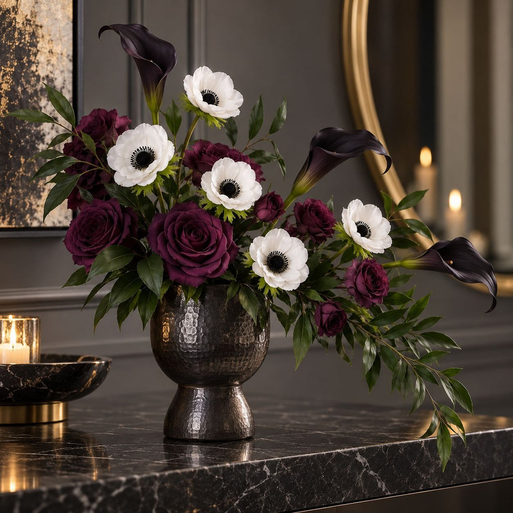A floral arrangement in a dark hammered metal urn vase, featuring burgundy roses, white anemones, and black calla lilies on a polished black marble console table.