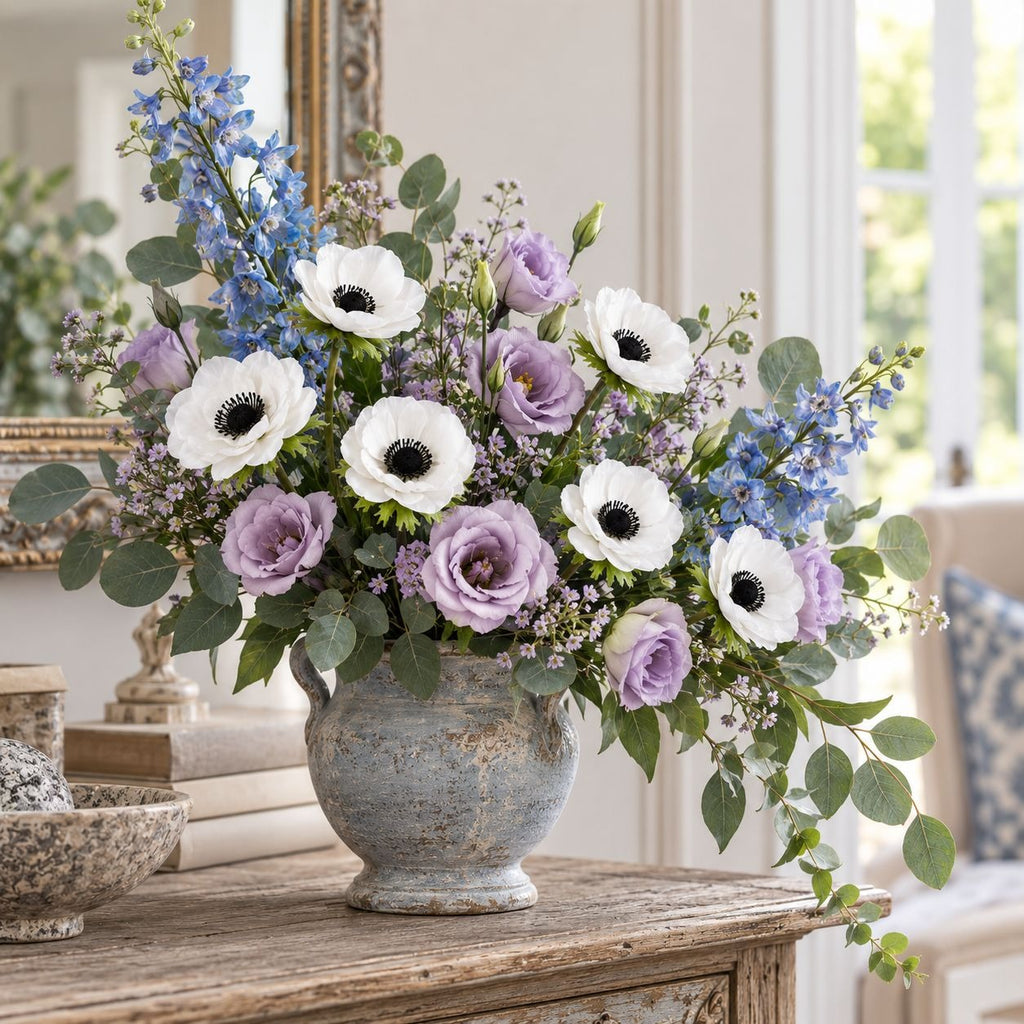 A floral arrangement in a distressed blue-gray ceramic vase, featuring white anemones, lavender roses, blue delphiniums, and green eucalyptus, displayed on a rustic wooden console table in a bright, elegant room.