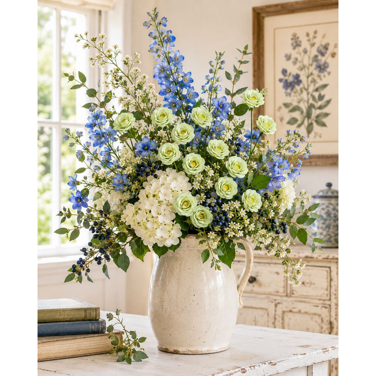 A large floral arrangement in a speckled cream ceramic pitcher vase, placed on a rustic white table in a bright, traditional room. The bouquet features light green roses, blue delphiniums, white hydrangeas, and green foliage.