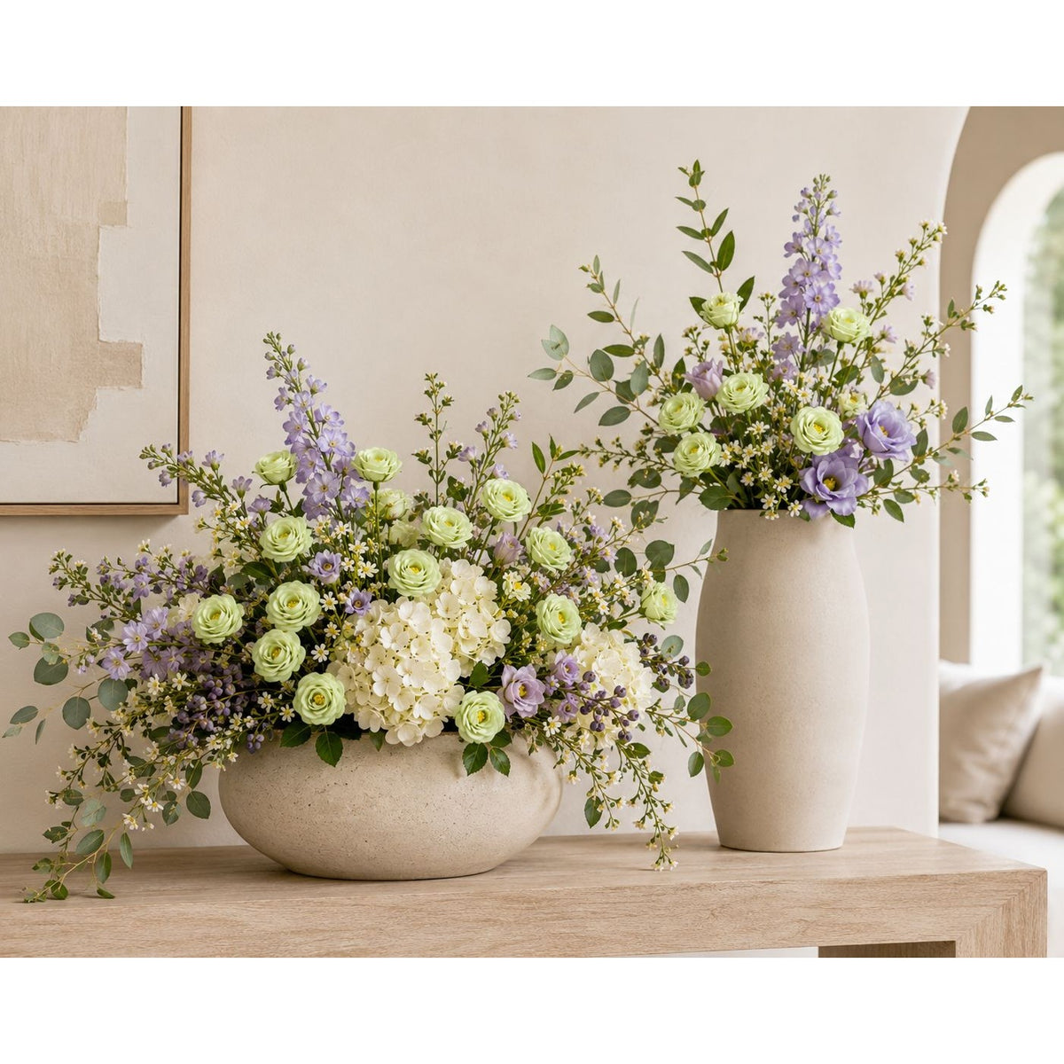 A styled floral arrangement in a round textured beige vase on a light wood table, next to a matching tall beige vase with a complementary flower arrangement, set against a warm beige wall.