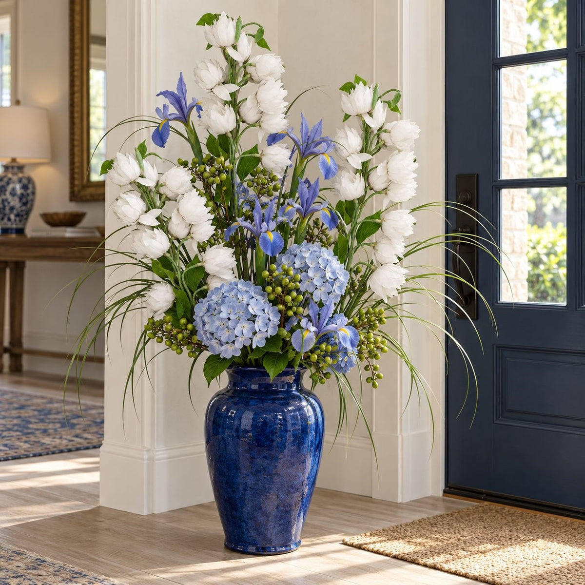 A large blue ceramic vase filled with a lush floral arrangement of white roses, blue hydrangeas, blue irises, and green foliage, placed in a bright entryway next to a dark blue door.