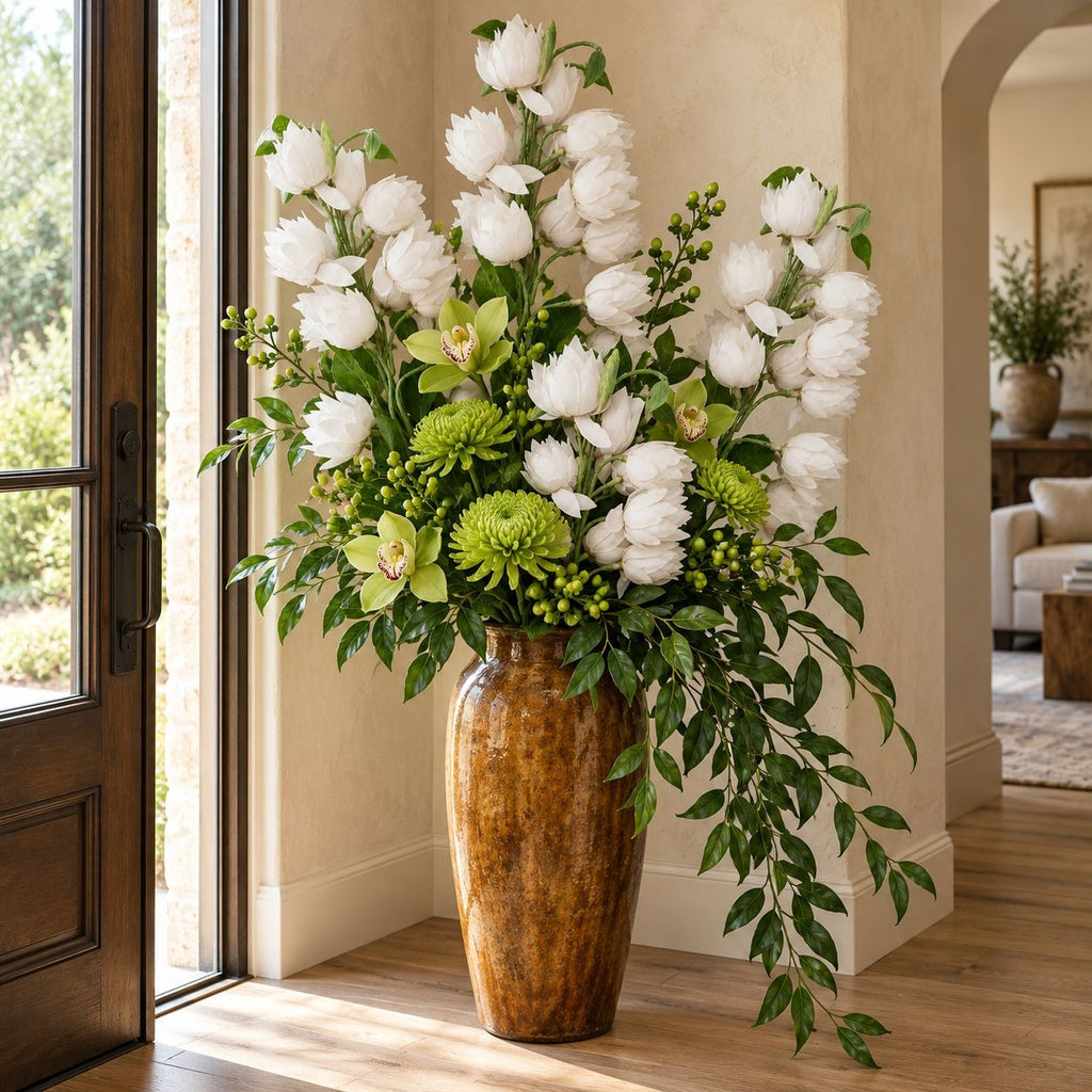 A large artificial floral arrangement in a tall brown ceramic vase, placed indoors near a glass door and living room background. The bouquet features white flowers, green foliage, and light green accents.