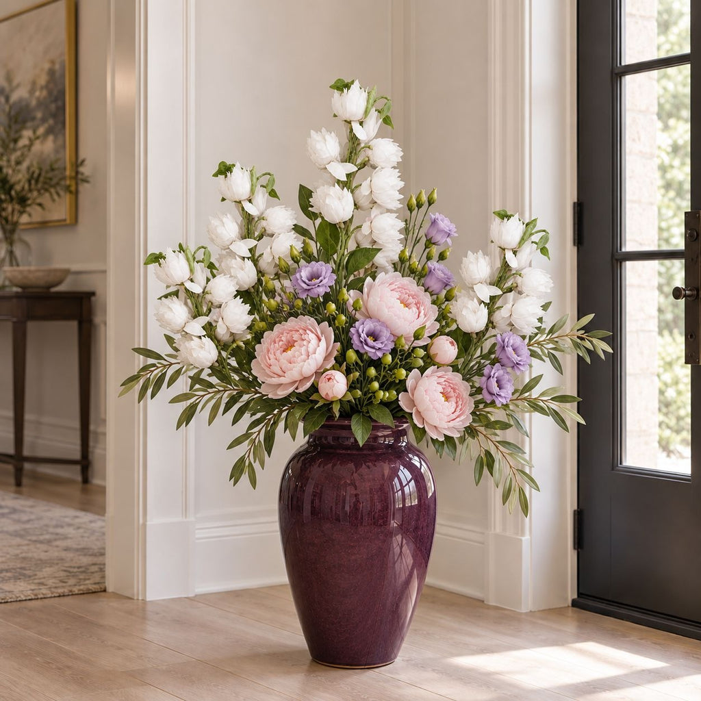 A large glossy dark purple ceramic vase filled with a lush floral arrangement of white ranunculus, pink peonies, and lavender sprigs, set in a bright, modern entryway.