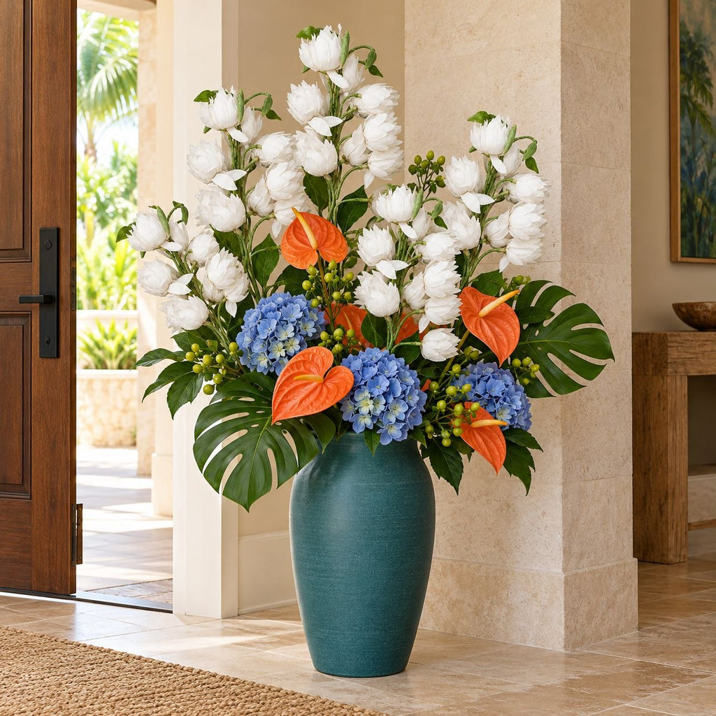 A large teal ceramic vase filled with a lush floral arrangement featuring white ranunculus, blue hydrangeas, orange anthuriums, and green monstera leaves, set in a bright indoor entryway.