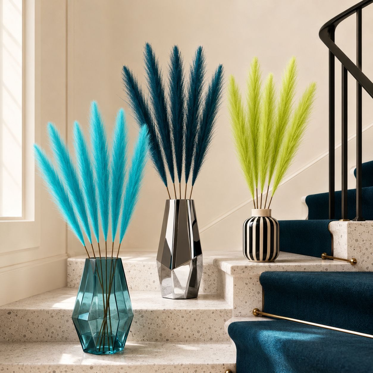 Three decorative pampas grass arrangements in different colored vases placed on indoor staircase steps. The left arrangement has bright turquoise grass in a translucent teal vase, the center has dark teal grass in a faceted silver vase, and the right has lime green grass in a black-and-white striped vase.