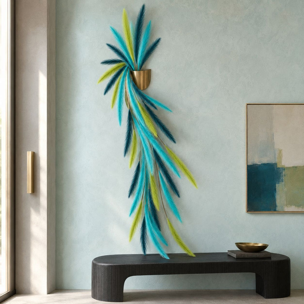 A decorative wall-mounted sculpture with long, flowing feather-like strands in turquoise, teal, and lime green, suspended from a small gold-toned cup holder, displayed in a styled interior setting above a dark bench.