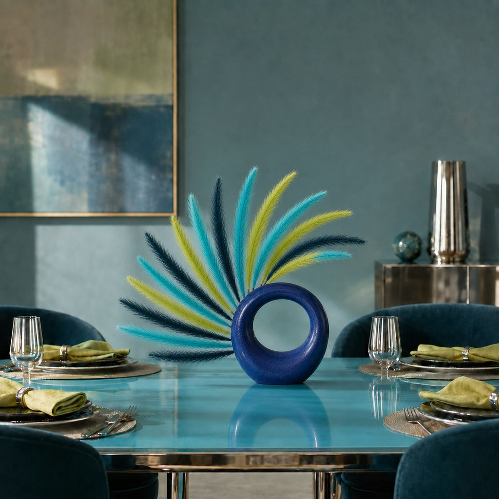 A decorative blue ring-shaped vase with a fan of blue, teal, and yellow feathers, displayed on a glossy turquoise dining table in a styled dining room setting.