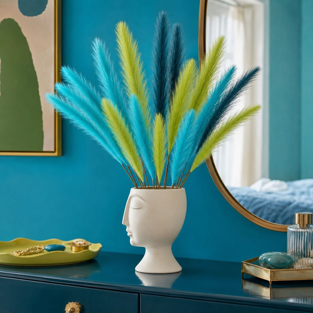 A decorative arrangement of fluffy pampas-style stems in bright blue, teal, and lime green, displayed in a white sculptural vase shaped like a side-profile human head, set on a dark blue dresser in a styled room with a mirror and wall art.