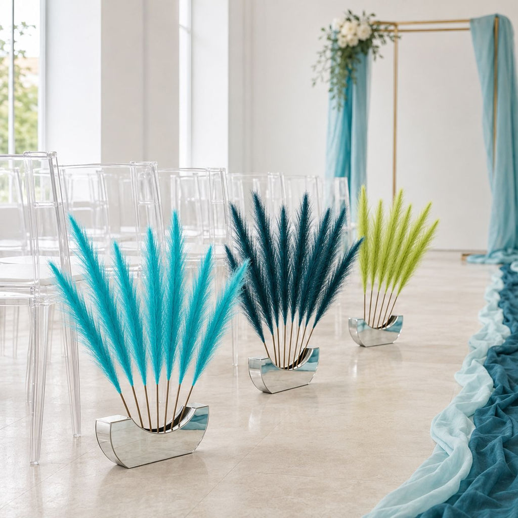 Three decorative pampas grass centerpieces in mirrored silver bases arranged on a light stone floor in a bright wedding-style venue. The left and center pieces are turquoise and deep teal-blue, while the right piece is lime green.