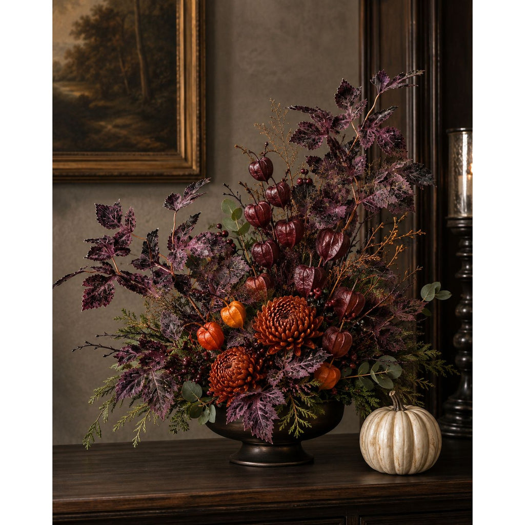 An autumn floral arrangement in a dark bowl vase, styled on a dark wooden mantel next to a small white pumpkin. The arrangement features deep burgundy and purple foliage, pinecones, small orange pumpkins, and berries, set against a dark wood panel and a muted brown wall.