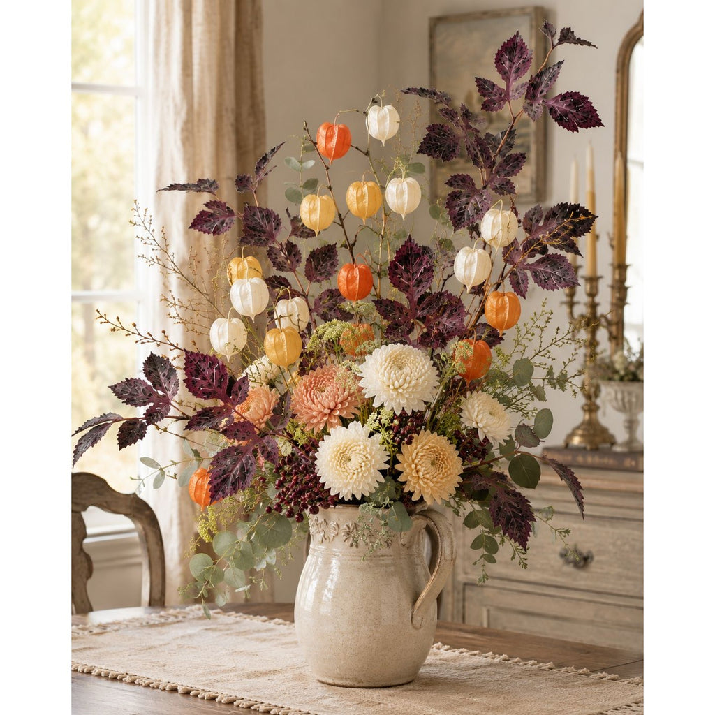 A large autumn floral arrangement in a rustic cream-colored ceramic pitcher vase, featuring orange pumpkins, white and cream chrysanthemums, and dark purple foliage, set on a table in a warm, vintage-style room.