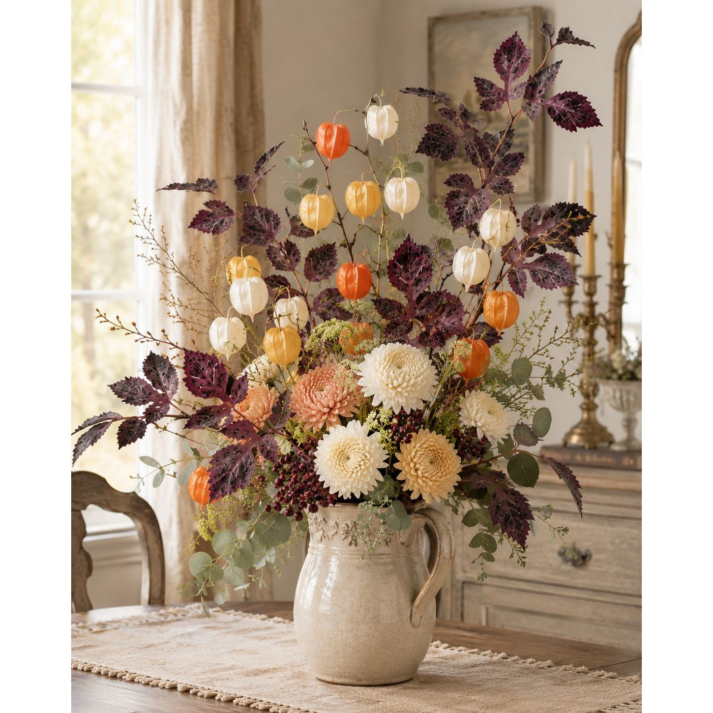 A large autumn floral arrangement in a rustic cream-colored ceramic pitcher vase, featuring orange pumpkins, white and cream chrysanthemums, and dark purple foliage, set on a table in a warm, vintage-style room.
