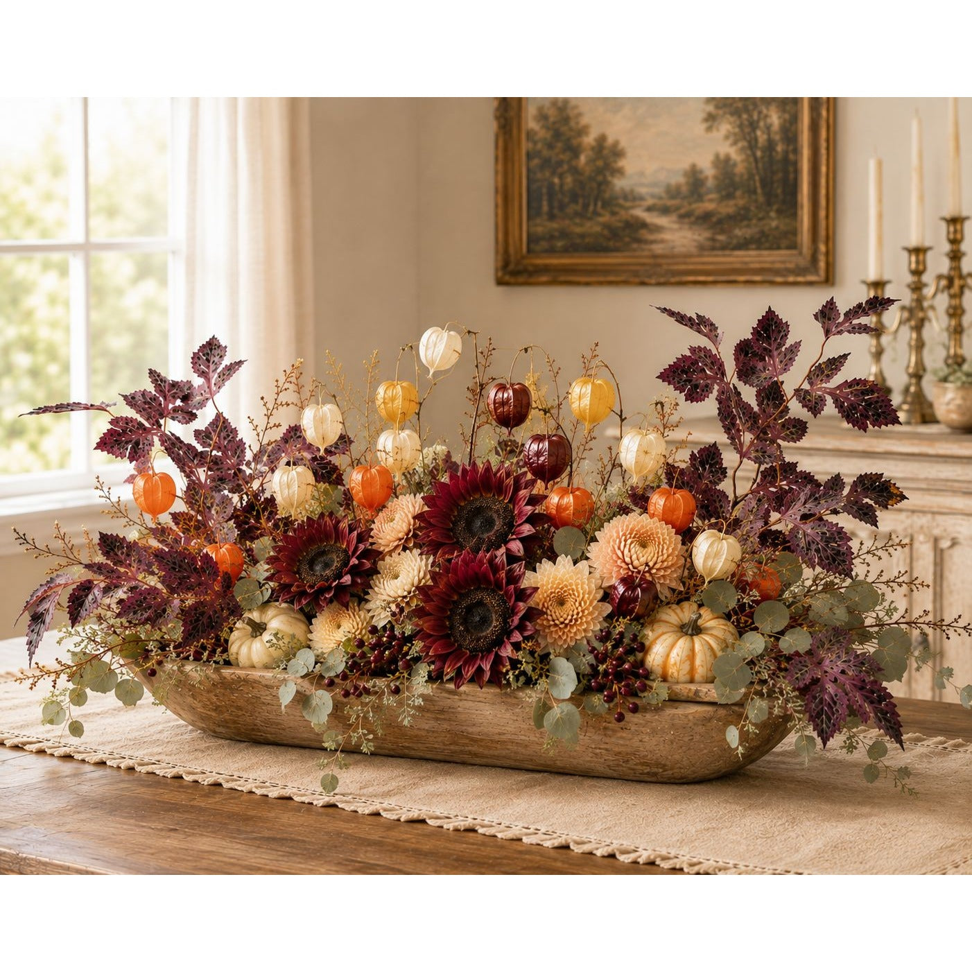 A rustic wooden oval tray filled with a fall floral arrangement featuring burgundy sunflowers, orange pumpkins, cream chrysanthemums, and dark purple foliage, displayed on a table in a warmly lit room.