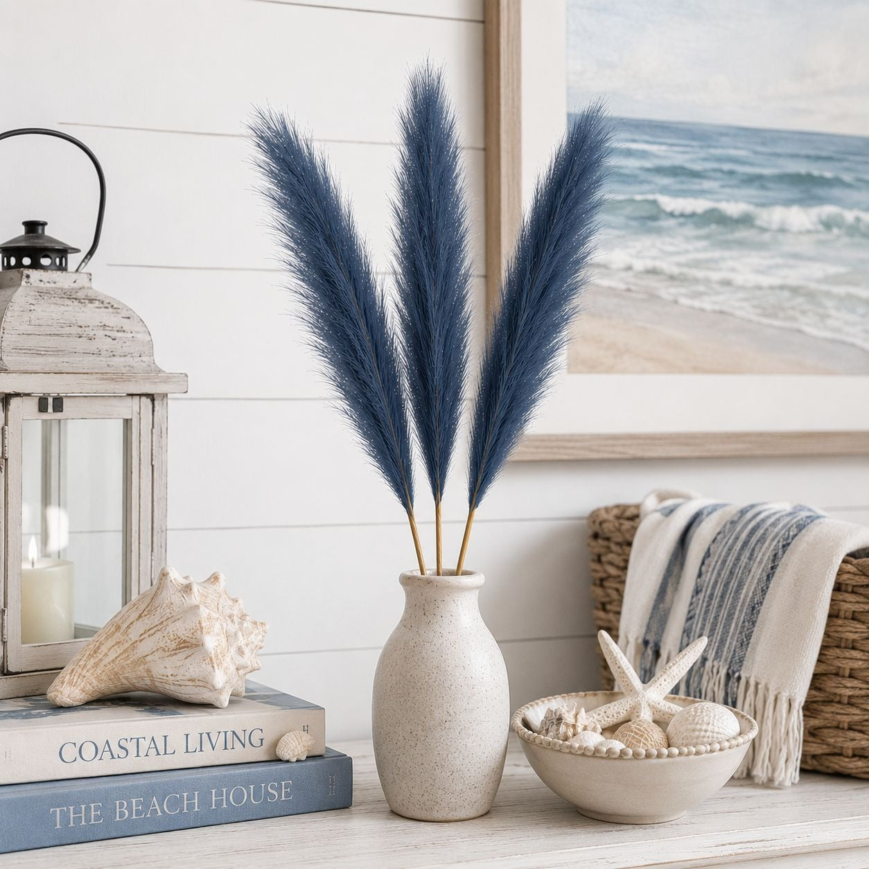 Decorative setup with pampas grass in a vase, coastal-themed books, lantern, and basket with starfish on a white surface.