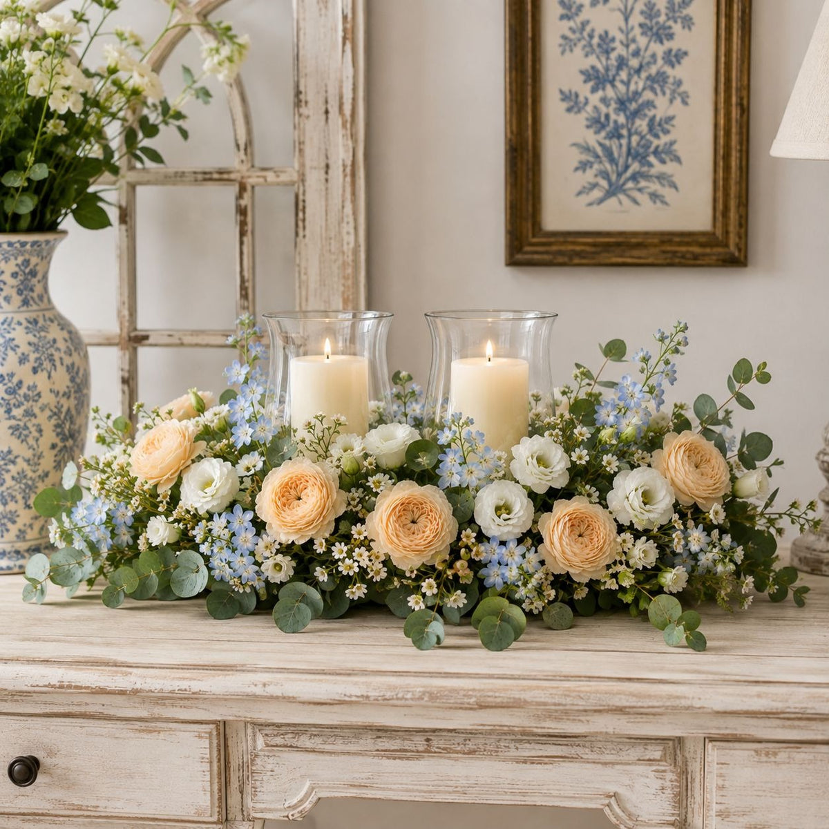 A floral centerpiece arrangement with two lit pillar candles in clear glass holders, placed on a distressed white wooden console table. The bouquet includes peach roses, white ranunculus, light blue flowers, and green eucalyptus leaves.