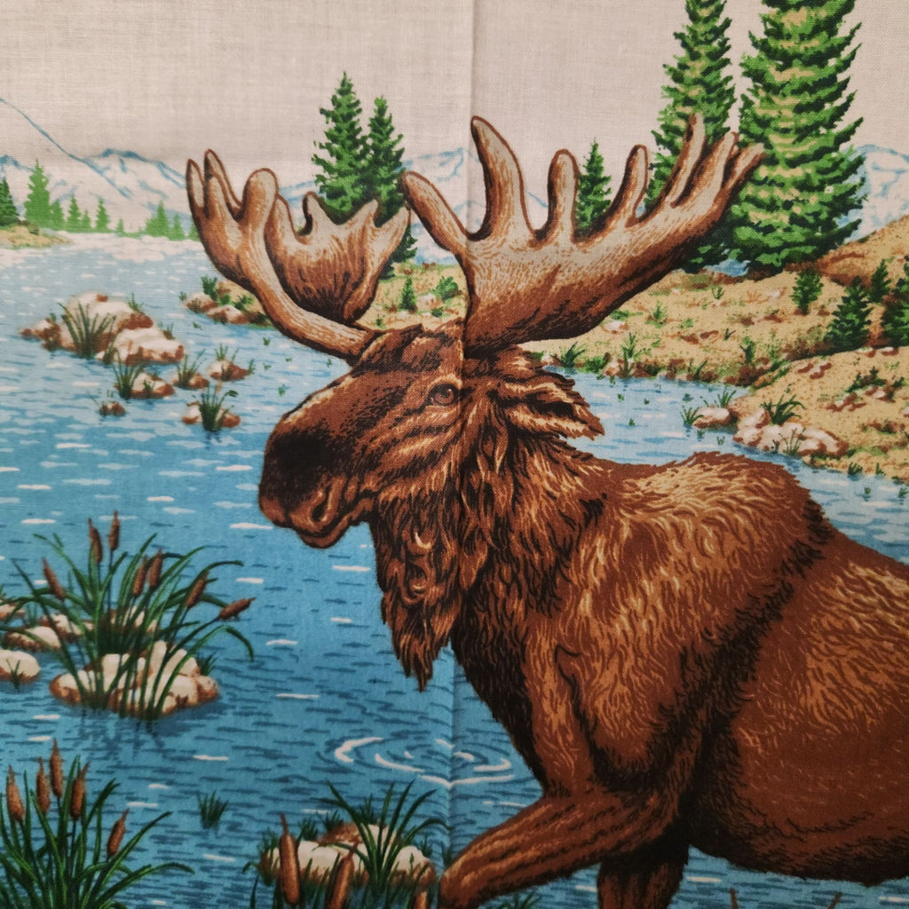 A close-up of a printed textile or fabric featuring a brown moose with large antlers standing in a blue river, with green pine trees, rocks, and mountains in the background.