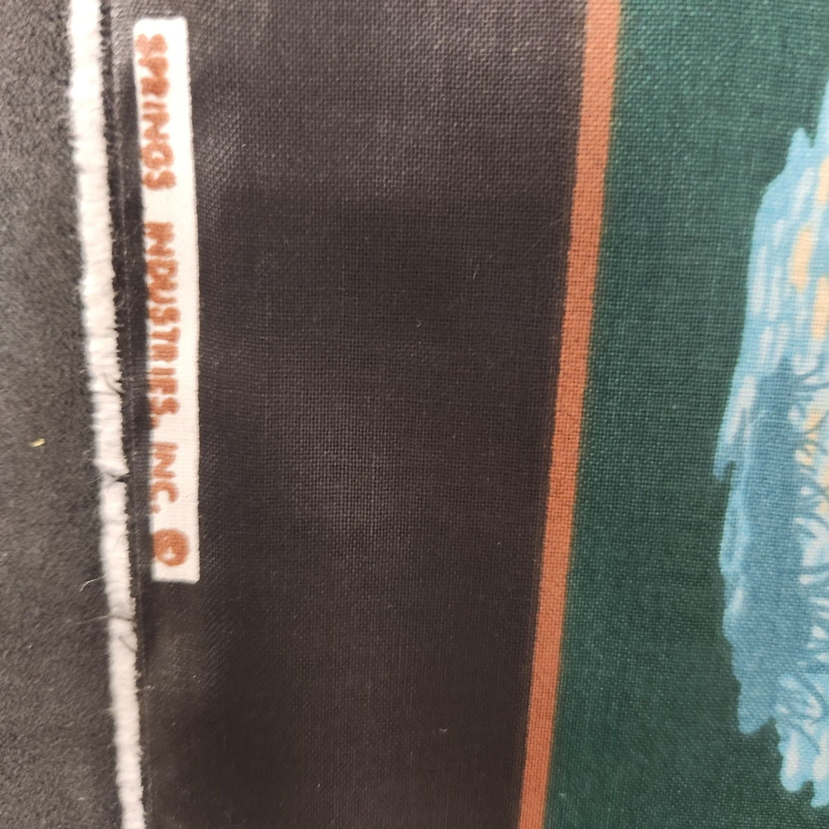 Close-up of a printed textile or fabric swatch showing a dark charcoal/black vertical band, a thin orange stripe, a green field, and part of a light blue graphic. A small sewn label reads 'SPRINGFIELD INDUSTRIES, INC.