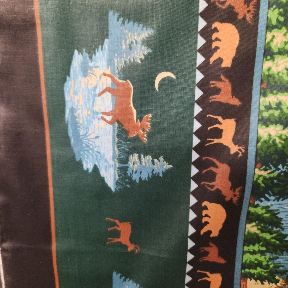 A close-up of a patterned fabricr featuring a moose in a snowy landscape, a crescent moon, and a vertical border with wildlife silhouettes on a dark background.